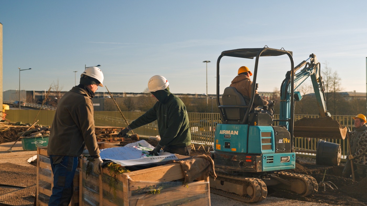 Team van Castelein Landscaping overlegt buiten naast bouwplannen met graafmachine op de achtergrond